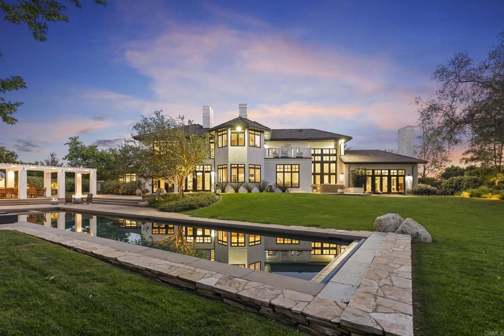 Twilight view of 7891 Muirfield Way: the rear elevation reflected in a 70-foot lap pool, illuminated interior windows under a dusk sky.